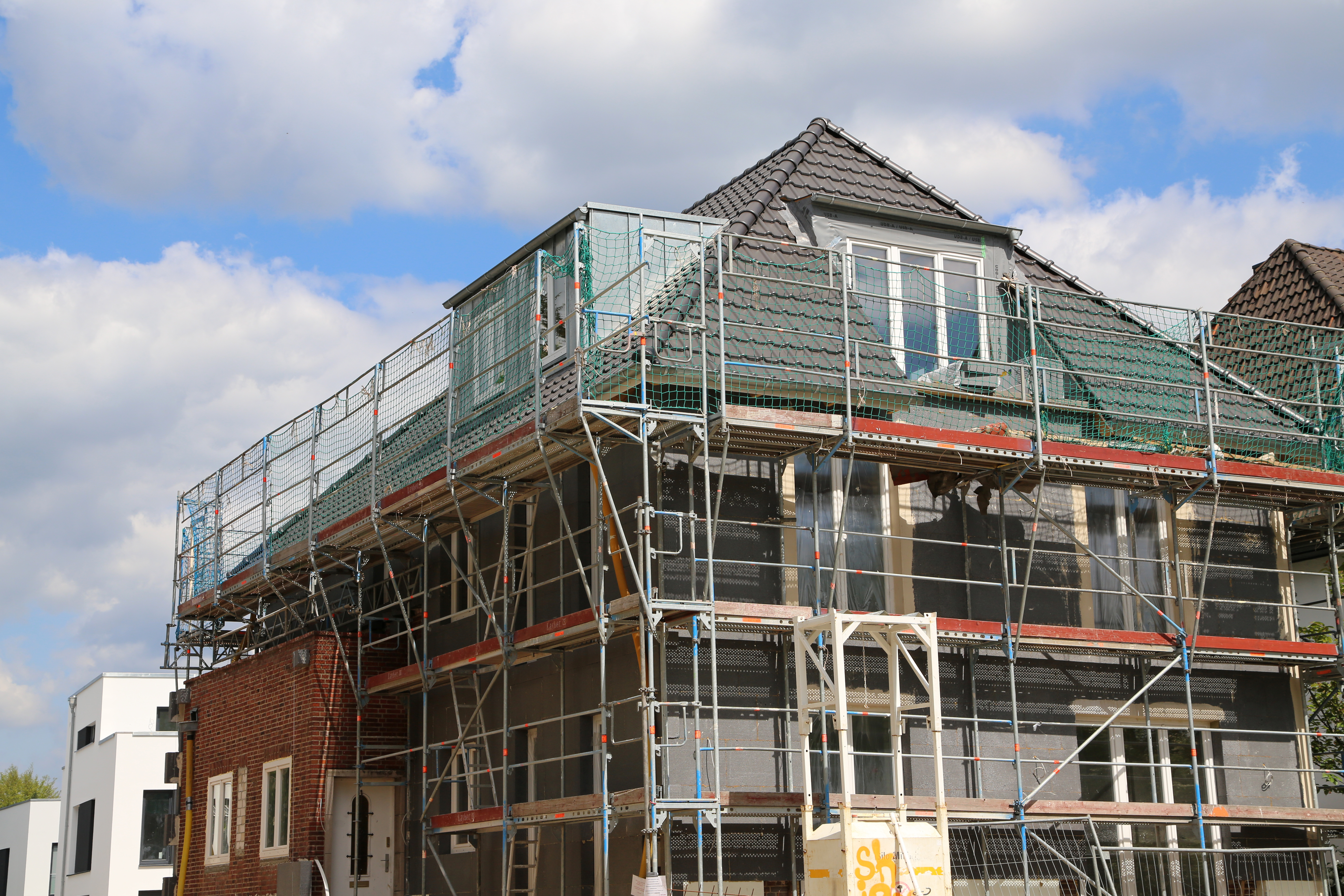 Sanierung-Haus-3 Ein mehrstöckiges Wohngebäude ist vollständig eingerüstet, während an Fassade und Dach im Rahmen einer Sanierung gearbeitet wird.